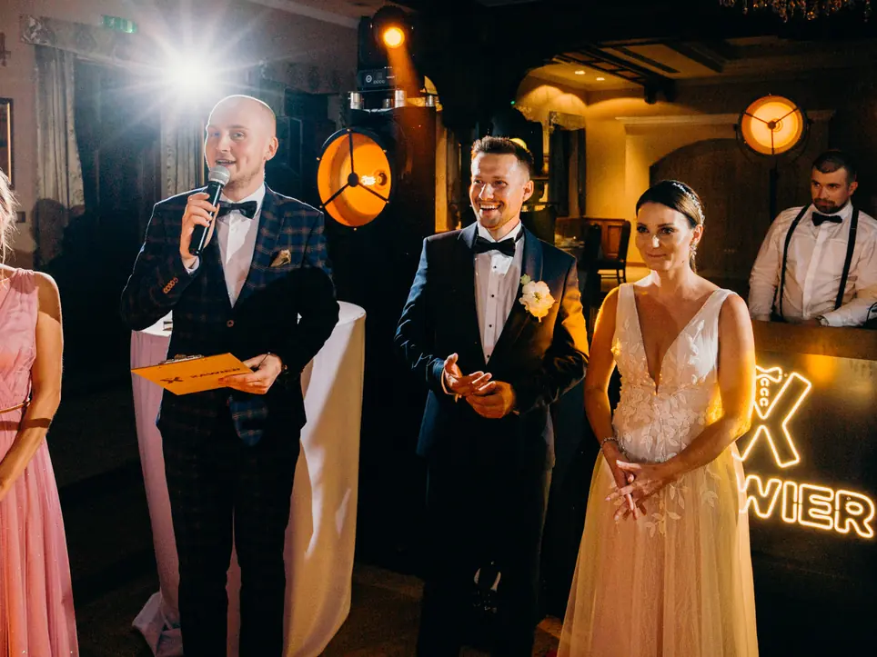 Wodzirej z mikrofonem obok Pary Młodej przy stanowisku z neonem